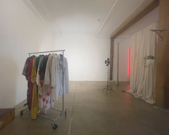 The image shows a spacious room with a clothing rack holding various garments, a light stand, and a curtain draped along one wall.