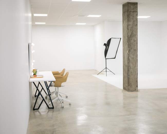 The image shows a spacious, minimalist studio featuring a white wall, polished concrete floor, two desks with chairs, and a lighting setup in the corner.