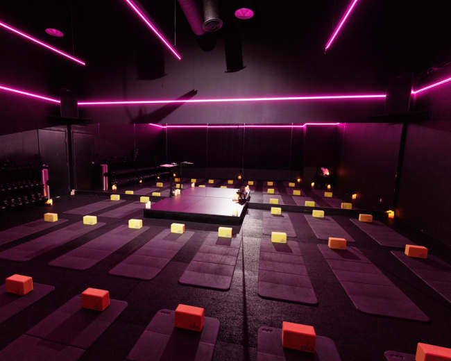 A dimly lit yoga studio features purple mats arranged neatly in rows, surrounded by orange yoga blocks and illuminated by pink accent lighting.