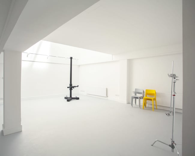 The image shows a spacious, minimalistic room with white walls, a skylight, a black lifting equipment, and two chairs—one gray and one yellow.
