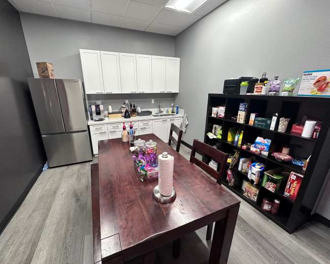 The image shows a kitchen area with a dining table, a refrigerator, white cabinets, and a shelf filled with various food items and supplies.