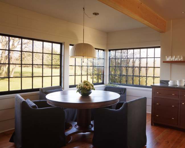 A round wooden dining table with four upholstered chairs is positioned beside large windows in a well-lit corner of a modern kitchen.
