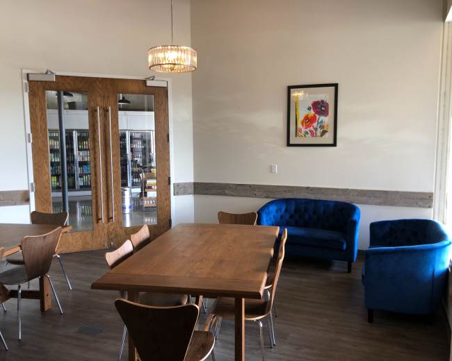 A well-lit cafe interior featuring a wooden table with four chairs, two blue velvet couches, and a framed floral artwork on the wall.