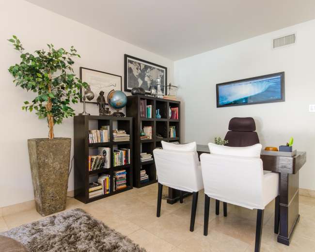The image shows a home office with a desk, two white chairs, a bookshelf filled with books, and a large potted plant in one corner.