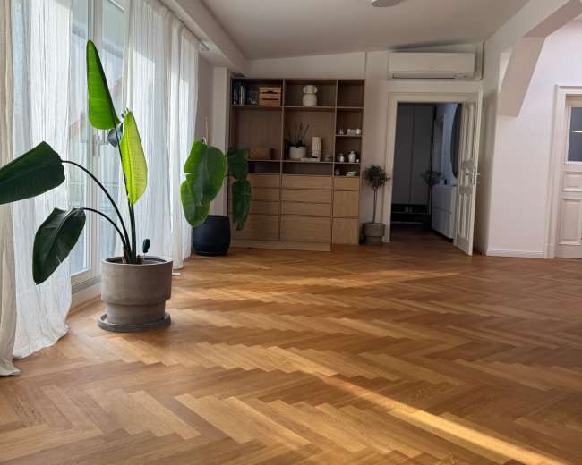 The image shows a spacious, bright room with a herringbone wood floor, large windows covered with sheer curtains, and a shelf filled with various items against one wall.