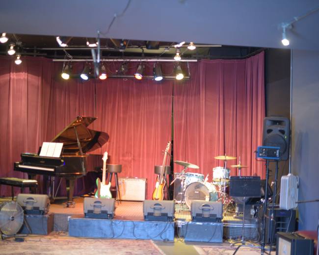 The image shows a stage setup with a grand piano, guitars, amplifiers, and a drum kit in front of a curtain backdrop.