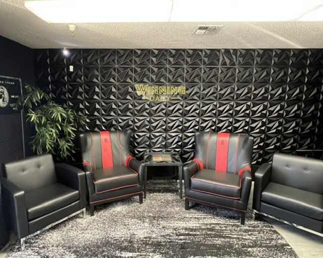 The image shows a modern lounge area featuring four black chairs, two of which have red accents, in front of a textured black wall with the words "WINNING DAILY" in gold lettering.