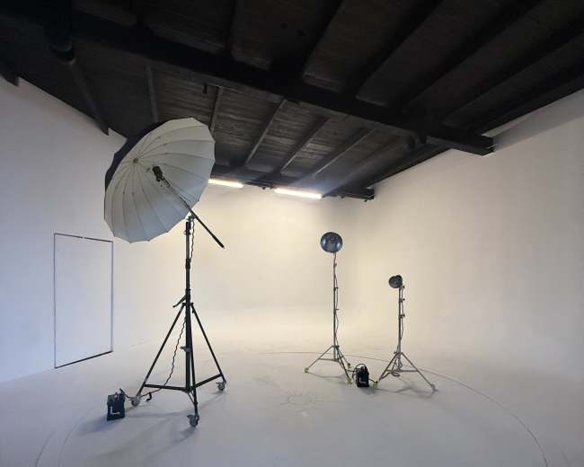 The image shows a minimalistic photo studio with a white backdrop, overhead lighting, and two light stands with an umbrella reflector.