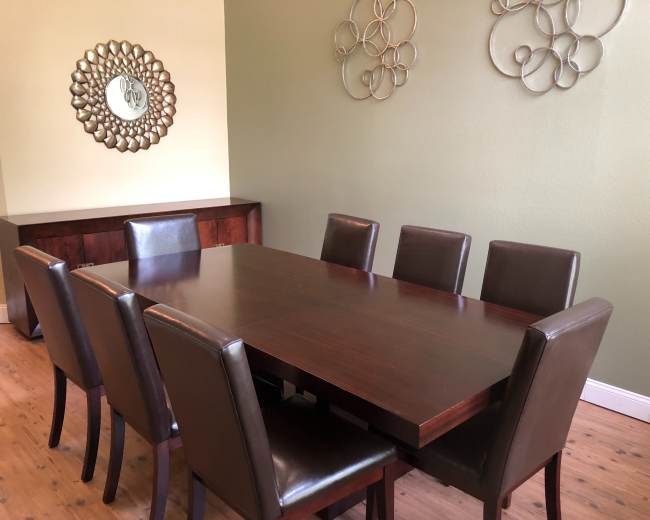 A wooden dining table with eight leather-upholstered chairs is positioned in a room with green walls and decorative metal wall art.