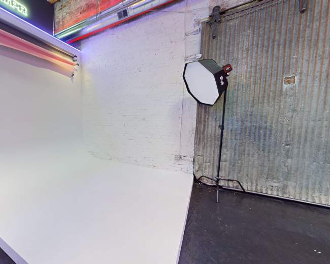 The image shows a photography studio with a seamless white backdrop and a softbox light positioned nearby against a textured wall.