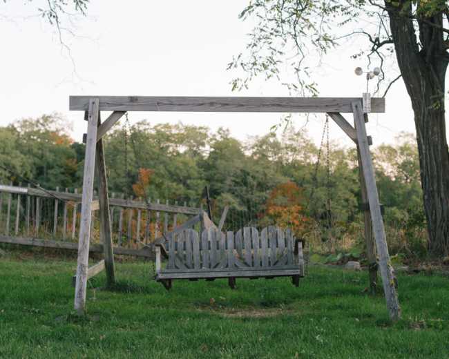 A wooden swing hangs from a frame in a grassy area, surrounded by trees.