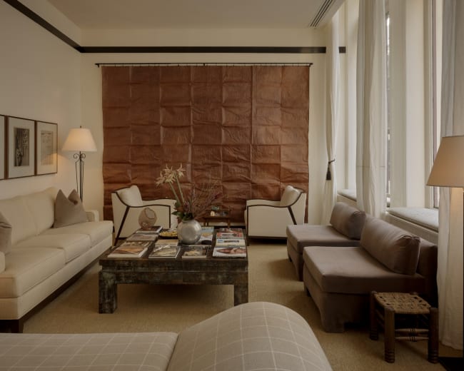 The image depicts a stylish living room featuring a beige sofa, two armchairs, a coffee table with magazines, and a textured brown wall hanging.