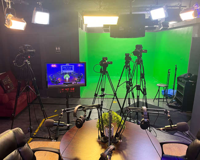 A television studio with multiple cameras, a green screen backdrop, and a central table equipped with several microphones.