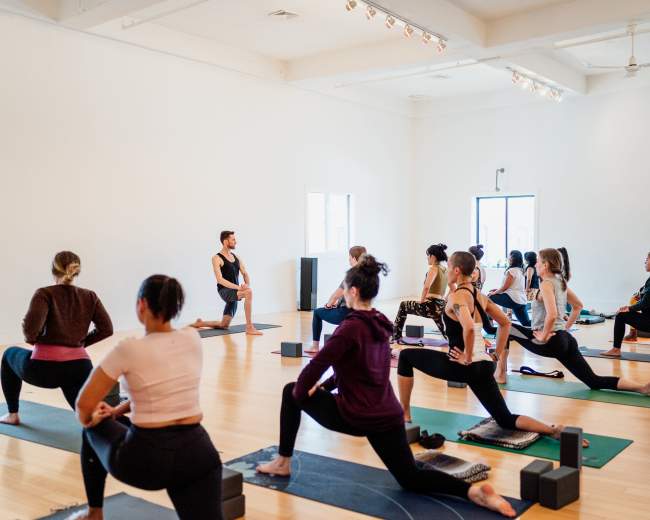 A yoga instructor guides a class of participants in a spacious studio, with students practicing lunges on their mats.