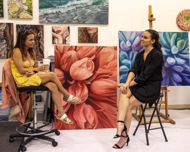 Two women are seated in an art studio surrounded by various colorful paintings, engaged in conversation.