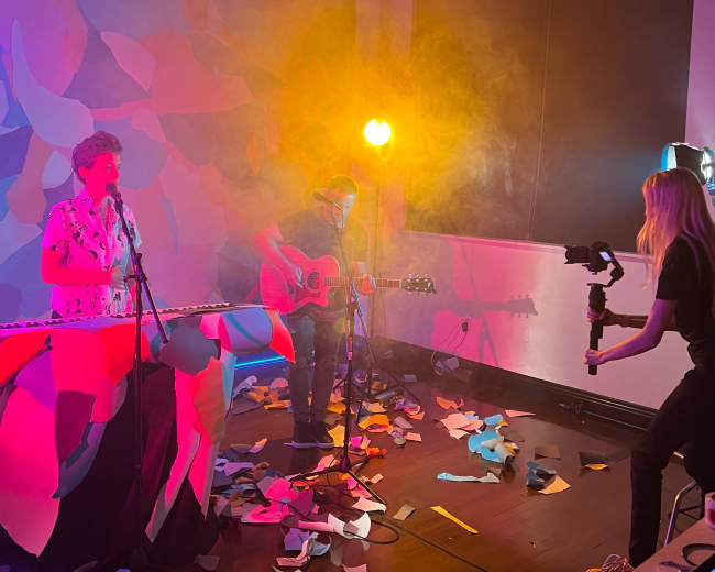 A musician plays guitar while a singer performs, surrounded by colorful paper scattered on the floor and an illuminated backdrop.
