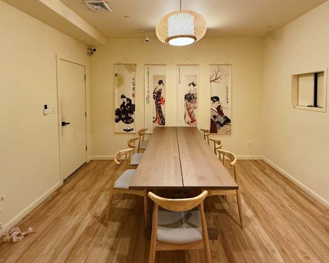 A long wooden table with six chairs is positioned in a softly lit room adorned with hanging scrolls featuring traditional Japanese images.