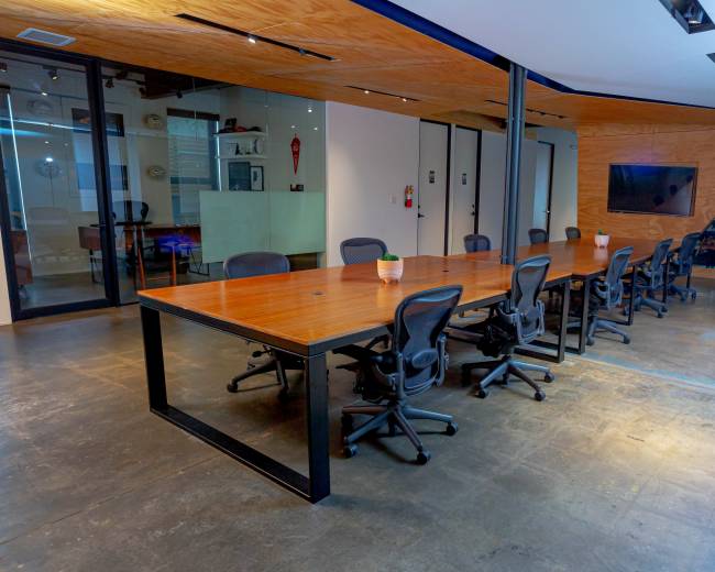 A long wooden conference table with black chairs is set in a modern meeting room with glass walls and a large screen.