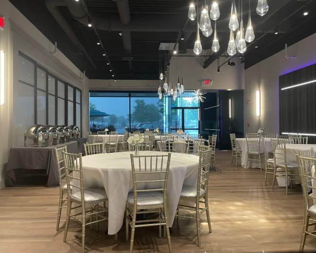 An elegantly set dining area with round tables, silver chairs, and a buffet setup, illuminated by modern pendant lights, and large windows revealing a cloudy exterior.