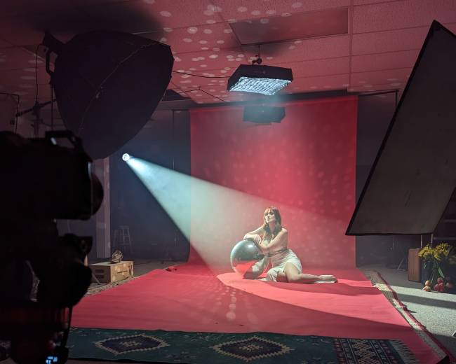 A model sits on a red backdrop in a photography studio, holding a globe while light beams illuminate the scene.