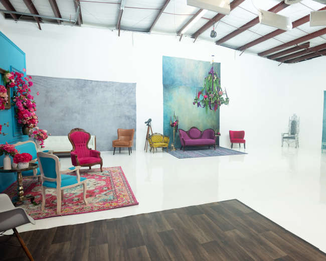 The image shows a bright and spacious photography studio featuring a variety of colorful furniture, including a purple couch, chairs, and decorative elements against a white backdrop.
