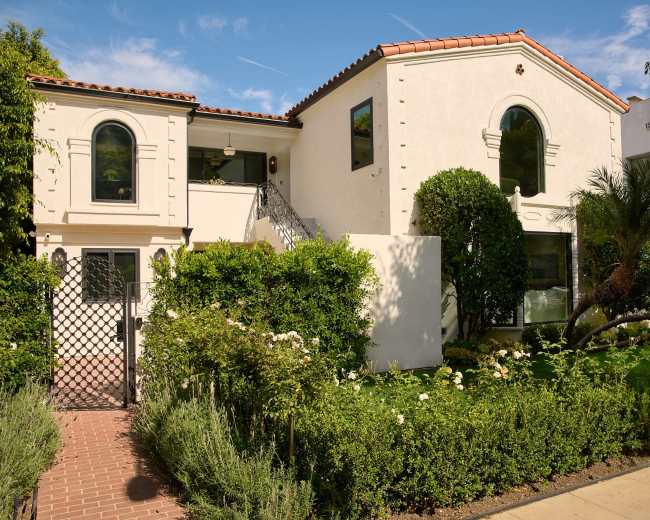 The image shows a two-story, white stucco house with arched windows and a tiled roof, surrounded by greenery and a well-maintained garden.