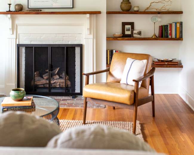 The image shows a living room with a fireplace, wooden shelves filled with books and decor, and a brown leather chair facing the fireplace.