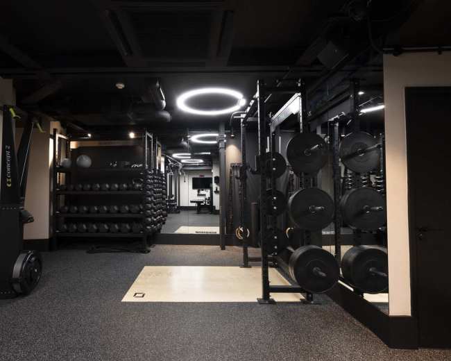 The image shows a modern gym interior featuring weightlifting equipment, free weights organized on racks, and a rubber flooring surface in a well-lit space.
