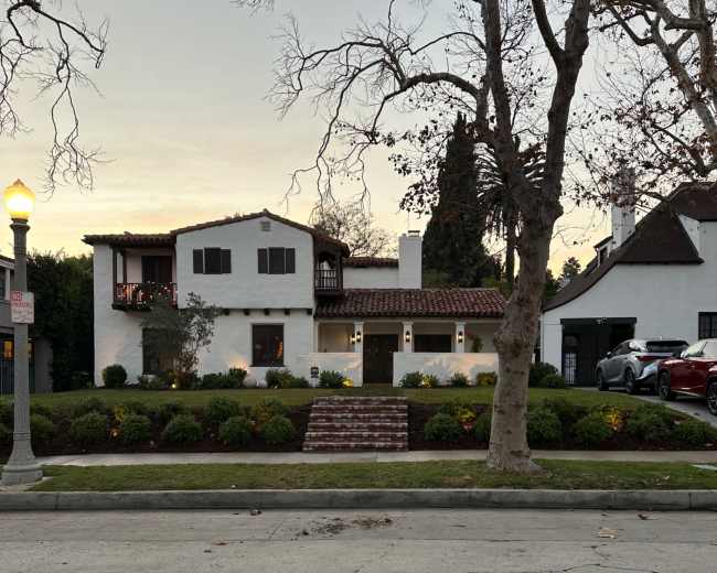 A white stucco house with a tiled roof and a front garden is situated along a street lined with trees.