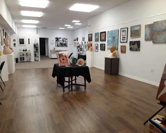 The image shows an art gallery interior with various artworks displayed on the walls and sculptures placed throughout the space.