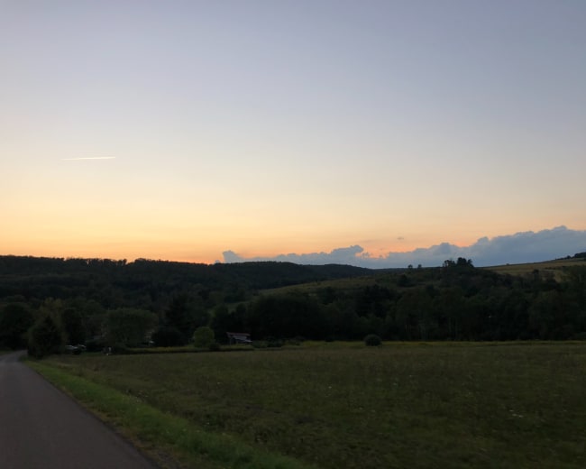 A paved road leads through a grassy field toward a horizon of rolling hills under a gradient sky at dusk.