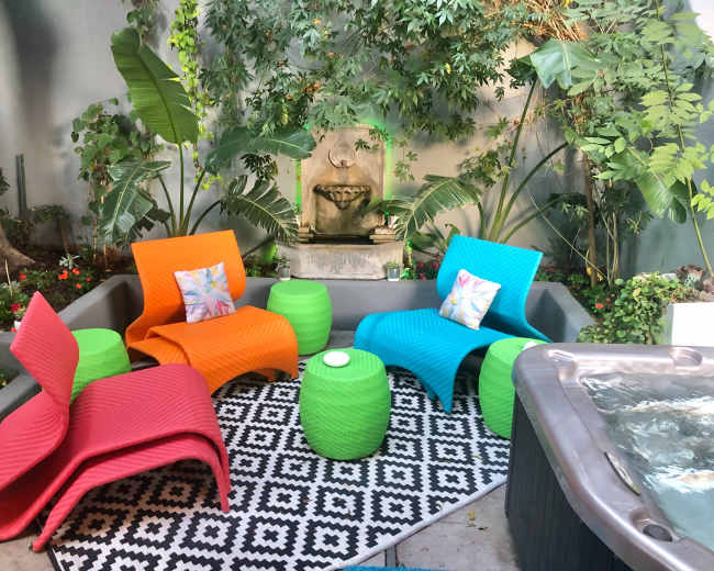 The image shows a vibrant outdoor seating area with colorful chairs, a patterned rug, potted plants, and a small fountain.