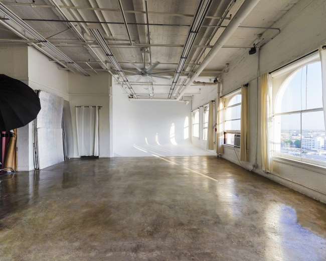 A spacious studio with large windows, concrete flooring, and a backdrop setup in the corner.