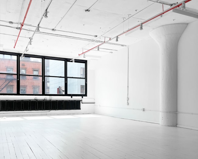 A spacious, minimalist room with white walls and floors, large windows, and exposed pipes.