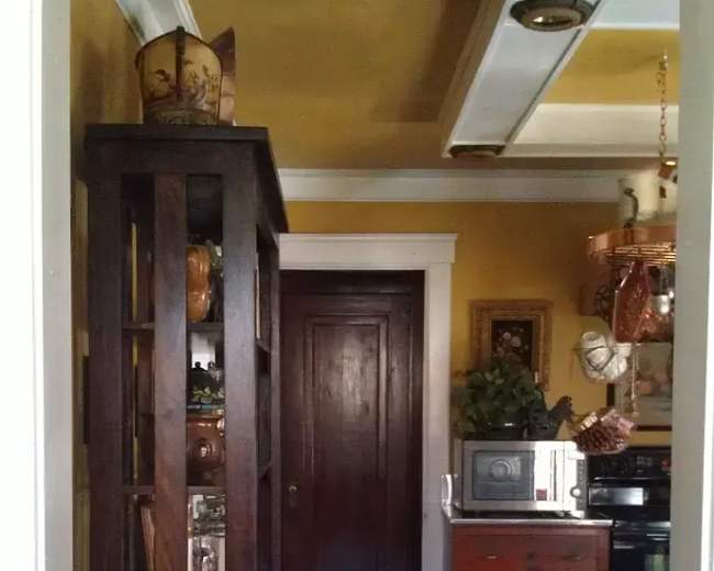 The image shows a narrow hallway leading to a door, with a wooden bookcase displaying various items on the left and a kitchenette area visible on the right.