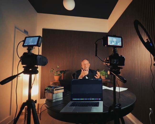 A man is sitting at a table in a dimly lit room, speaking into a microphone while being recorded by cameras on tripods.