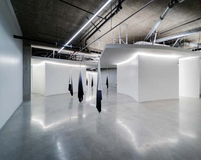 The image shows an interior space with smooth, curved walls, polished concrete flooring, and black and white hanging fabric installations.