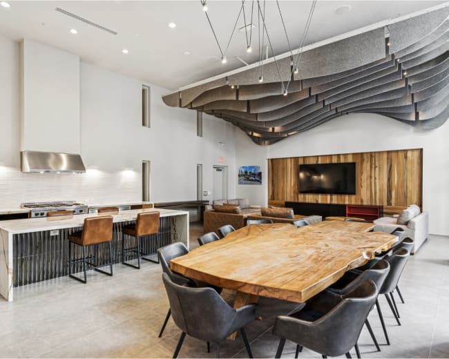A modern communal space features a large wooden dining table surrounded by chairs, a kitchen area on one side, and a textured ceiling design above.