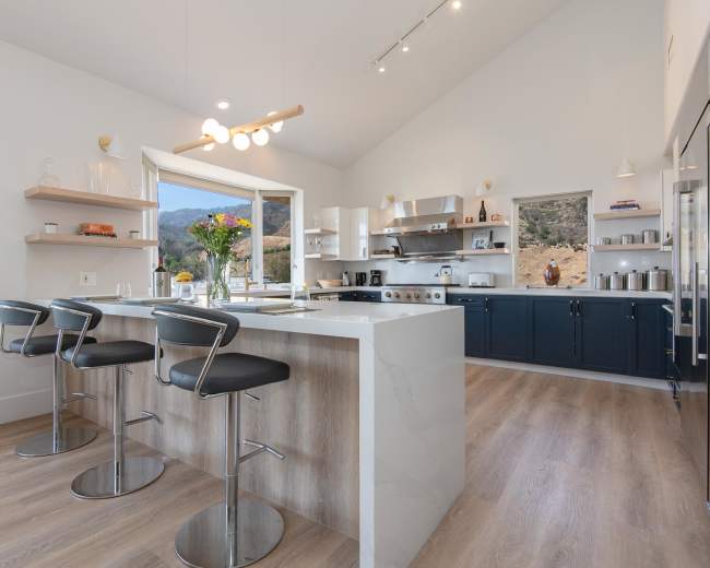 The image shows a modern kitchen featuring a large island with three bar stools, sleek cabinetry, and large windows that provide natural light.