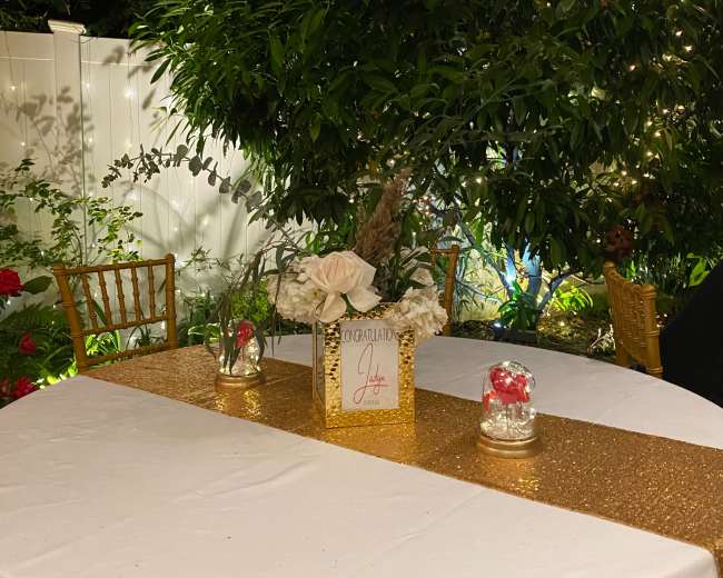 A neatly arranged table with a gold table runner, a floral centerpiece, and decorative candles is set in a garden surrounded by greenery and flowers.