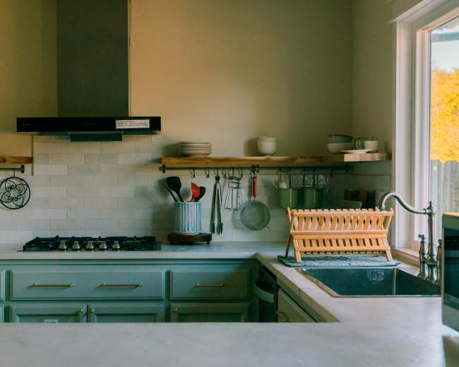 The image shows a modern kitchen with a gas stove, wooden shelves holding dishes, and a dish rack near the sink.