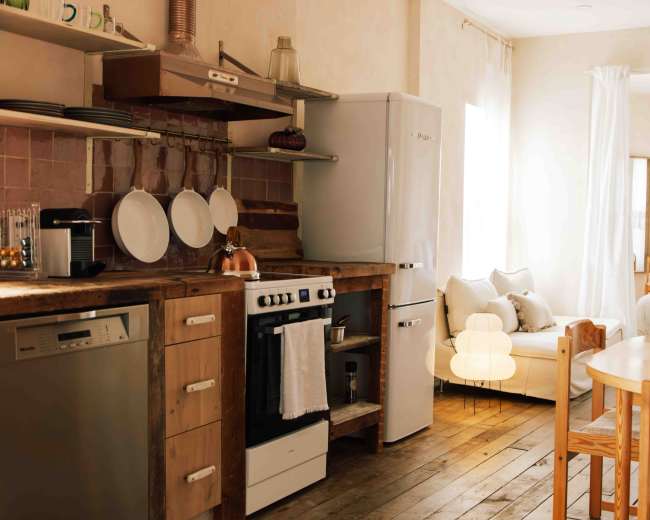 The image shows a modern kitchen area with a wooden counter, a white refrigerator, a stove, and a dining table, all set in a bright, airy space.