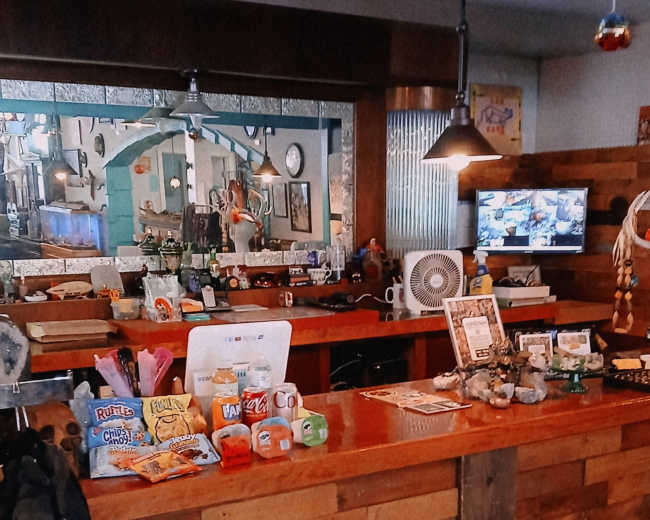 The image shows a cozy bar area with a wooden counter, displaying various snacks and drinks, and decorated with unique items, including a championship belt.