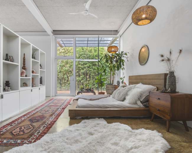 A modern bedroom featuring a low wooden bed, a decorative rug, and shelves displaying various objects, with large windows allowing natural light and views of greenery outside.