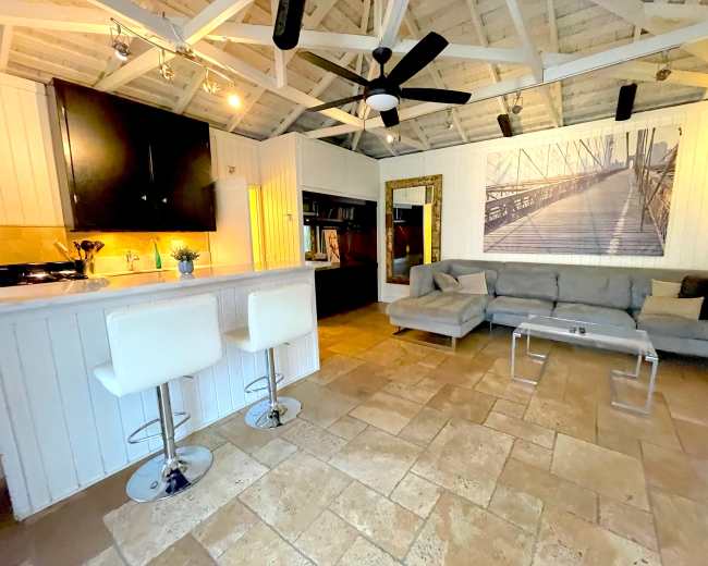 The image shows a spacious interior with a kitchen area featuring bar stools, a living area with a gray sofa, and a large wall art piece depicting a bridge.