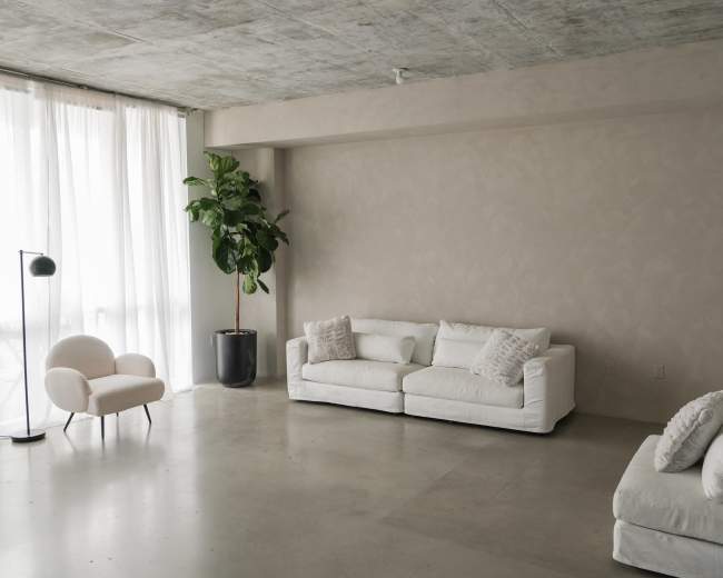 A spacious living room features white sofas, a small chair, a floor lamp, and a potted plant, with large windows allowing natural light to enter.