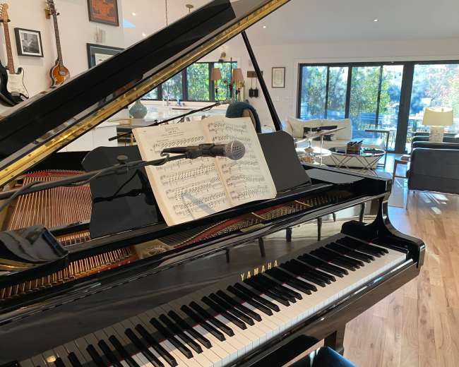 A grand piano with sheet music and a microphone is positioned in a bright room featuring large windows and a view of outdoor greenery.