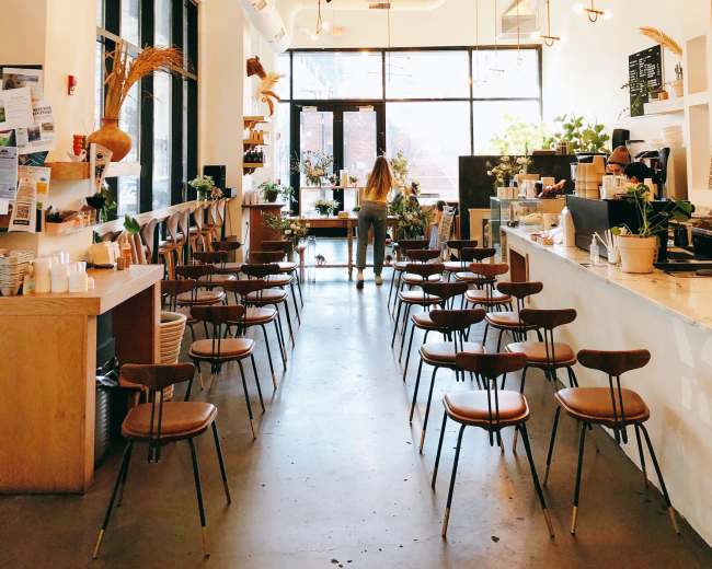 The image shows a spacious, modern café interior with wooden chairs arranged in rows and large windows letting in natural light.