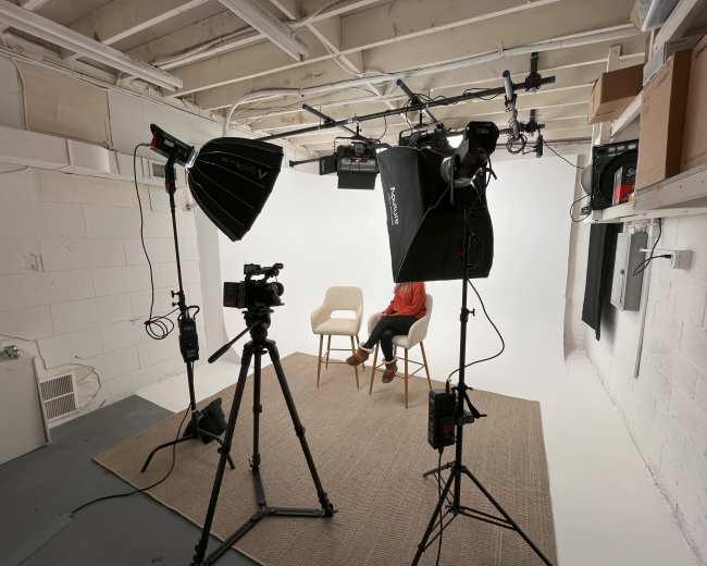 A studio setup features two chairs on a rug, with multiple lights and cameras positioned to capture a seated individual against a white backdrop.