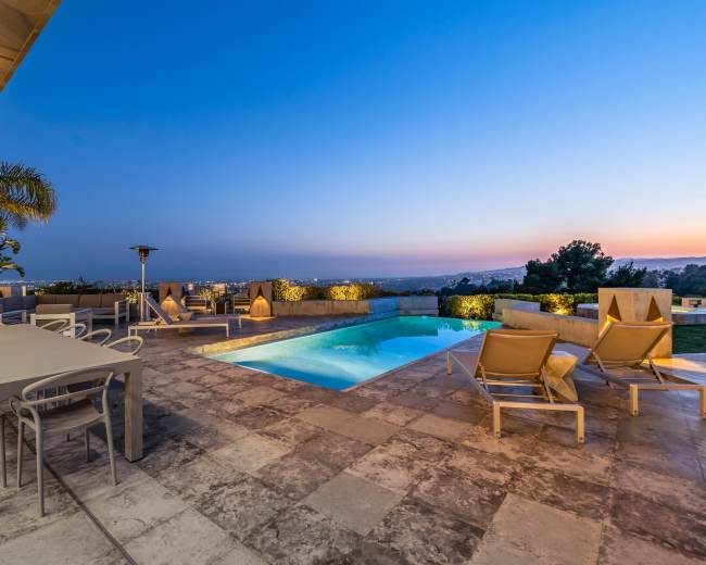 A modern outdoor patio features a swimming pool, lounge chairs, a dining table, and a view of the city skyline at dusk.